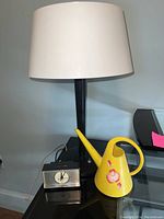 Full view of the lamp with white shade, vintage General Electric clock, and yellow watering can on black surface