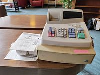 Front view of Sharp XE-A101 cash register on a wooden table with paper rolls and instruction manuals nearby.