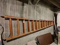 Full length view of the wooden ladder showing rungs and side rails lying horizontally against a concrete wall in a basement or storage area.