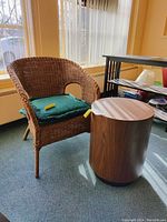 Wicker accent chair with green cushion next to round wooden side table on carpeted floor in a well-lit room.