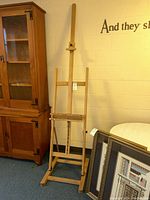 Full view of the Windsor and Newton wooden easel standing upright with adjustable vertical and horizontal supports, placed beside wooden cabinet and picture frames.