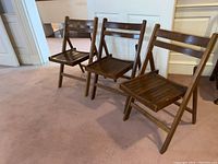 View of three wooden vintage folding chairs arranged in a row showing front and side angles, set on carpet flooring in front of white doors and walls. Medium brown finish wood with slatted seat and backrest visible.