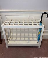 Frontal view of white painted wooden umbrella stand with one colorful umbrella placed inside. Stand has a lattice style top and shelf bottom showing some dirt and marks.