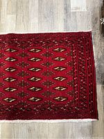 Close-up image showing the intricate repeating gul flower pattern and rug border in red shades with cream, black, and brown details on wooden floor.