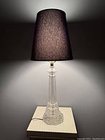 Full view of the lamp on a textured table, showing the black lampshade and clear crystal glass base illuminated from behind.