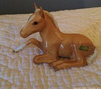 Palomino horse figurine laying down on quilted fabric, showing overall front-left-side view.