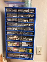 Front view of blue wall-mounted organizer with multiple clear drawers filled with assorted small hardware and fasteners