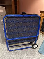 Full folded bed showing blue metal frame and wire spring base, mattress folded inside frame and wheels on bottom for mobility.