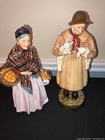 Front view of two Royal Doulton figurines: The Orange Lady holding baskets of oranges and Lambing Time holding two lambs
