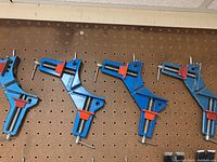 Four blue and red corner woodworking clamps hung on a pegboard wall, showing overall condition and color.