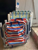 Four colorful folding beach chairs stacked against a wall. Two chairs have Tommy Bahama branding on the fabric backrest with colorful stripes and aluminum frames.