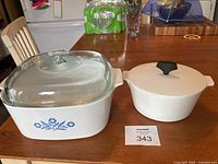 Photo showing both Corning Ware pieces side-by-side on a table, the larger casserole with floral design and glass lid and smaller buffet server with plain white lid and black knob.