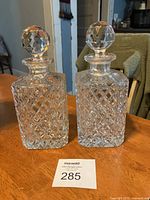Two clear crystal decanters with diamond pattern cut and fitted round stoppers on a wooden surface.