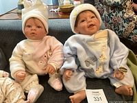 Pair of vinyl dolls seated on a couch, showing front view of boy in blue and girl in pink fleece pajamas with hats.