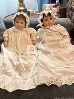 Two vinyl collector dolls in white detailed gowns and bonnets seated on a sofa.