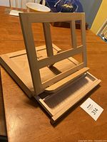 Angled front-side view showing wooden easel with frame and bottom support ledge, drawer partially open.