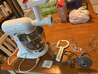 Wide shot showing KitchenAid Artisan stand mixer in white color with glass bowl and meat grinder attachment installed. Several accessories laid out on the table including wire whisk, flat paddle, dough hook, plastic flat attachment, and clear plastic pouring shield.