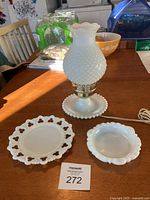 Full view of the electric milk glass hurricane lamp and two types of milk glass trinket dishes/plates on wooden table.