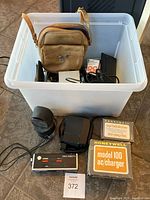 Entire lot in a plastic storage tub showing multiple camera accessories, lenses, a brown leather shoulder strap bag, and boxed Honeywell AC charger.