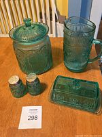 Photo showing teal glass cookie jar with knob lid, beverage jug with handle, and salt and pepper shakers with metal tops on wooden surface.