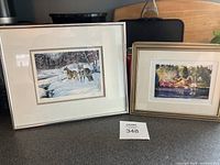 Two framed art prints on a dark surface: left print depicts wolves in snow, right print shows boat house and fishing trawlers scene.