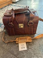 Full view of the vintage brown leather camera case showing overall exterior, top handles, shoulder strap laid out in front, and metal clasp.
