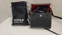 Black leather case with red interior containing Kurt Muller binoculars next to a black Fujifilm Instax Mini branded drawstring pouch.