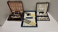 Three boxed cutlery sets arranged side by side: two dark cases with silver-plated fish forks and one blue case with stainless steel knives featuring Bakelite handles.