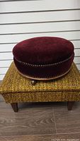 Round burgundy velvet stool with brass nailhead trim on small castors on floor.