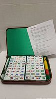 Open zippered rectangular brown case with green fabric lining containing Mahjong tiles, dice, chips, and instruction sheet.
