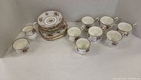 Full tea set view showing 7 side plates stacked, 7 cups arranged, 8 saucers, and two additional bowls (cream and sugar) in white background.