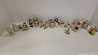 Wide shot showing all 16 small porcelain figurines arranged in a row on white surface with plain cornered background.