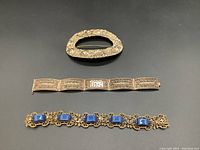 Three vintage jewelry pieces on black background: large curved floral stamped pin, silver filigree bracelet missing stones, and a blue stone bracelet with gold-tone filigree metal.