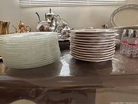 Two stacks of side plates on a table: the left stack consists of clear glass plates with a subtle textured pattern, the right stack consists of cream-colored octagonal porcelain plates decorated with a grapevine motif.