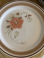 Single ceramic plate showing hand-painted style orange flower with brown leaves and stems on cream background, brown rim