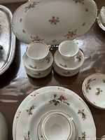 Stack of porcelain teacups and saucers with floral designs and gold rim, large oval floral platter visible behind.
