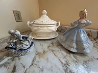 Photo showing the soup bowl tureen with ladle and spillage tray along with the two ceramic figurines on a marble surface