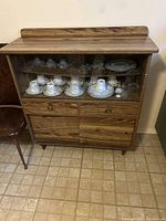 Full front view of the wood-veneer buffet cabinet with glass door showing the contents inside and lower drawers closed.