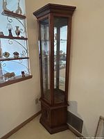 Full view of tall, narrow wood veneer display cabinet with glass door and shelves in corner of room.