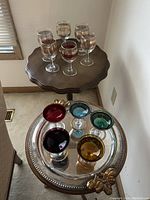 First photo of two sets of glasses; 5 tall clear stemmed glasses on a wooden table and 6 colored short stemmed glasses on a silver decorative tray