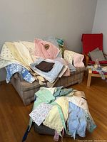 Various vintage towels and aprons with pastel colors and floral patterns, some with crochet borders, arranged on a sofa and ottoman.