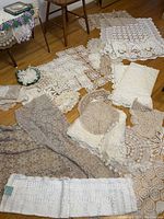 Overview of various crochet linens, including doilies and tablecloths in cream, beige, and white tones arranged on floor around furniture legs.