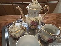 Porcelain teapot, creamer, sugar bowl on metal serving tray reflecting items