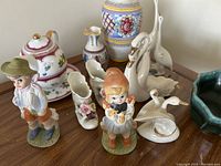 Photo showing boy and girl figurines, pair of ceramic boot planters with floral embellishments, swan creamer, teapot with floral design on saucer, small decorative pitcher, and large vase with colorful floral pattern.