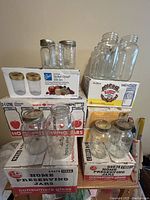 Vintage glass mason jars in assorted sizes stacked on original boxes labeled 'Home Preserving Jars' and 'Lards'. Some jars have metal screw lids, others are lidless.