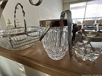 Two glass ice buckets and one glass salad bowl with metal handles placed on a wooden surface with natural light coming through a window.