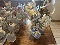 Photo showing 6 white espresso cups with matching saucers arranged on a metal rack along with 7 hanging vintage patterned mugs.