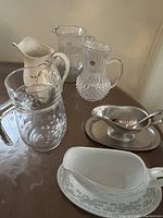Four pitchers and two gravy boats arranged on a surface showing clear glass, ribbed crystal, and ceramic items with gold accents