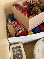 Photo showing two stacked cardboard boxes filled with assorted used Christmas ornaments including baubles, stocking, and pine cone shaped ornaments.