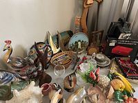 Overview of miscellaneous ceramic figurines, decorative plates, flowers, glassware, cash box, and kitchen utensils.